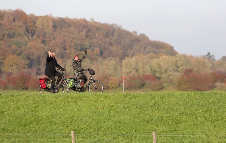 Burgemeester Kottelenberg en zijn vrouw fietsen op de dijk en zwaaien naar de camera.