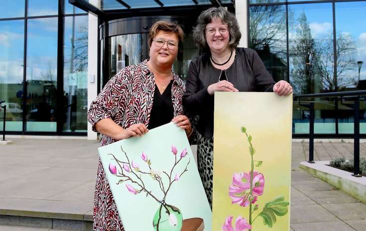 Wethouder Herma van Dijkhuizen en kunstenaar Roelien Everts poseren samen met twee schilderijen voor het gemeentehuis.