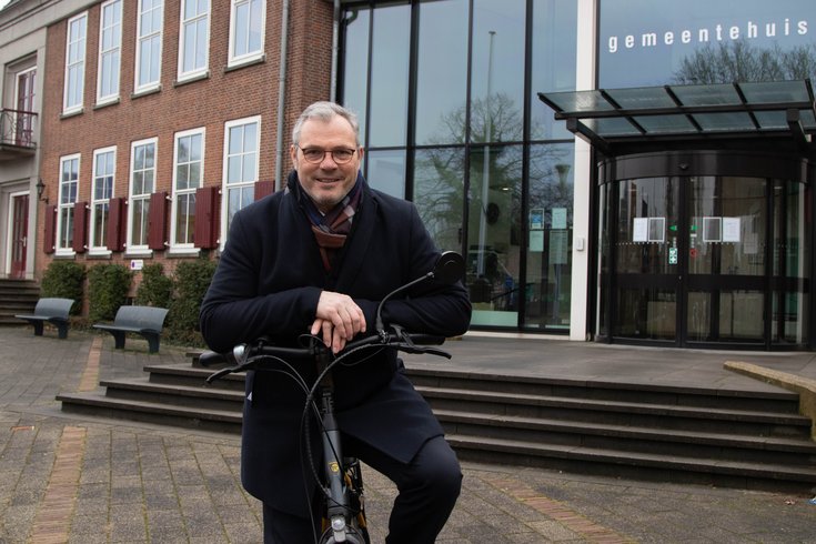 Burgemeester Jan Kottelenberg lacht naar de camera terwijl hij op het stuur van zijn fiets leunt en voor het gemeentehuis staat.